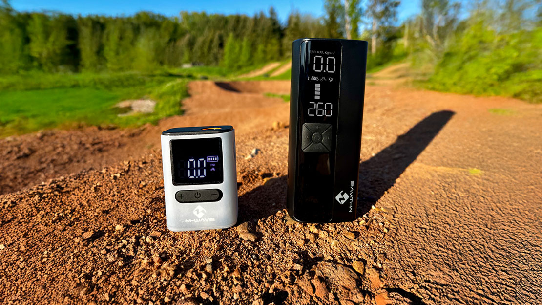 M-WAVE brand digital air pumps or tire pressure gauges on a dirt road or trail. Trees and a clear blue sky can be seen in the background. The devices stand on a loamy surface, one silver-colored and more compact, the other black and larger with an extended display.